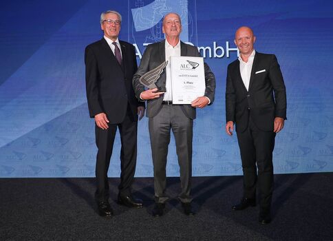 Auf einer Bühne stehen drei Herren in Anzügen; der mittlere Preisträger hält eine Urkunde und eine Glas-Trophäe, während die beiden anderen ihn beglückwünschen. Blauer Hintergrund mit Mustern.