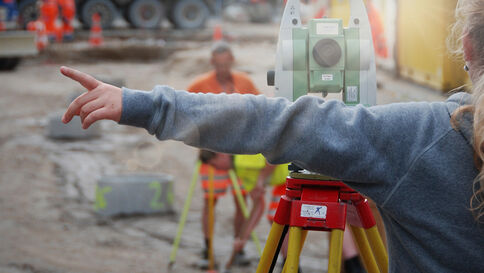 Im Vordergrund steht ein grünes Vermessungsgerät auf einem Stativ; eine Person in grauem Hoodie zeigt nach links. Im Hintergrund arbeiten Bauarbeiter in orange Sicherheitswesten, umgeben von Absperrungen auf einer Baustelle.