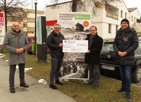 Vier Männer stehen draußen vor einer Werbetafel und halten gemeinsam einen großen Spendenscheck; es wirkt wie eine Scheckübergabe im Freien, vor Gebäuden und geparkten Autos.