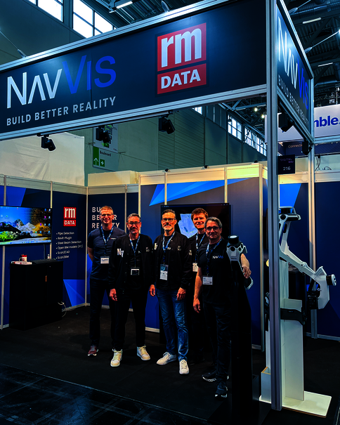 Gruppe von fünf Personen mit Namensschildern posiert vor dem NAVVis-Messestand mit RM Data-Logo. Großes Banner oben: NAVVis Build Better Reality. Hinter ihnen weitere Stände, links Monitor, rechts ein weißes Messgerät.