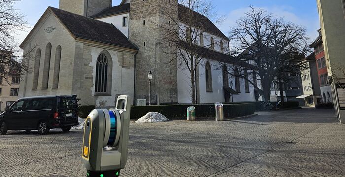 Eine Kirche mit zwei hohen Türmen und roten Spitzen steht in einer kleinen Stadt; die Türme tragen Uhren. Der Platz davor ist gepflastert, von Bäumen und Gebäuden umgeben.