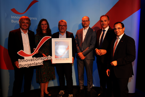 Sechs Personen stehen auf einer Bühne; eine Person in der Mitte hält ein gerahmtes Zertifikat, eine weitere ein Cut-out-Schild mit der Aufschrift 'Innovationspreis Burgenland'. Im Hintergrund ist ein Banner mit dem Logo zu sehen.