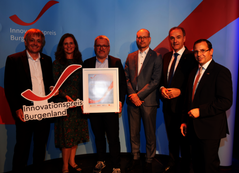 Sechs Personen stehen auf einer Bühne; eine Person in der Mitte hält ein gerahmtes Zertifikat, eine weitere ein Cut-out-Schild mit der Aufschrift 'Innovationspreis Burgenland'. Im Hintergrund ist ein Banner mit dem Logo zu sehen.