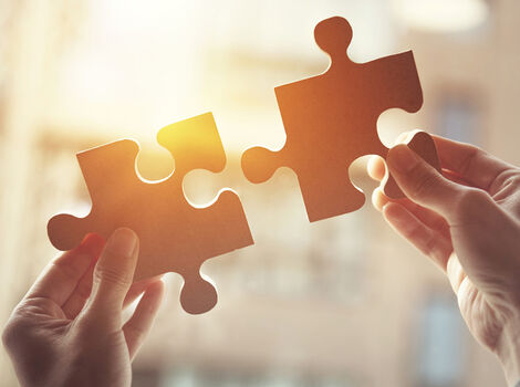 Zwei Hände halten zueinander passende Puzzle-Stücke, die kurz davor sind, ineinander zu greifen. Im warmen Licht im Hintergrund entsteht ein Symbol für Zusammenarbeit.