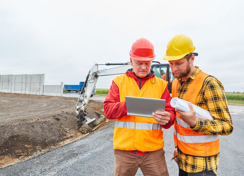 Zwei Bauarbeiter auf einer Baustelle prüfen Pläne: einer hält ein Tablet, der andere einen Bauplan. Sie tragen orange Sicherheitswesten und Helme (rot und gelb); im Hintergrund sieht man einen Bagger und Erdarbeiten.