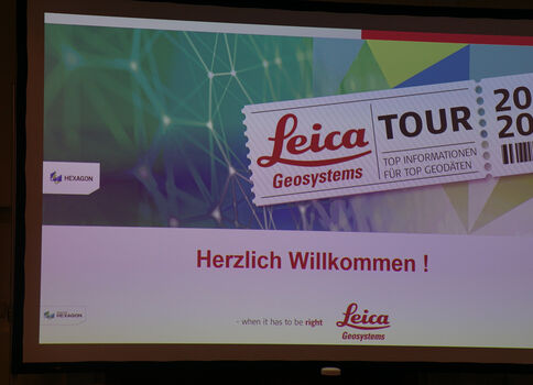 Begrüßungsfolie auf einem Bildschirm: Buntes geometrisches Hintergrundmuster; ein Banner mit 'Leica TOUR 2020' und der Begrüßung 'Herzlich Willkommen!'; unten Leica Geosystems-Logo und weitere Logos.