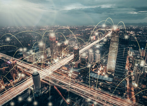 Nachtaufnahme einer modernen Großstadt mit hellen Straßen und Hochhäusern; darüber ziehen leuchtende Lichtlinien und Knotenpunkte, die ein digitales Netzwerk und die Vernetzung der Stadt symbolisieren.