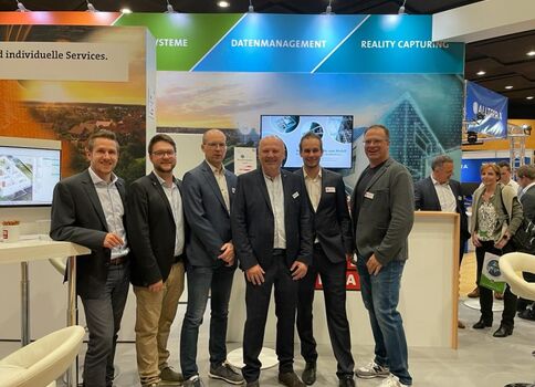 Sieben Männer in Anzügen posieren lächelnd vor einem Messestand. Der Hintergrund zeigt Banner mit Worten wie Systeme, Datenmanagement und Reality Capture; weitere Besucher und Messeinfrastruktur sind sichtbar.