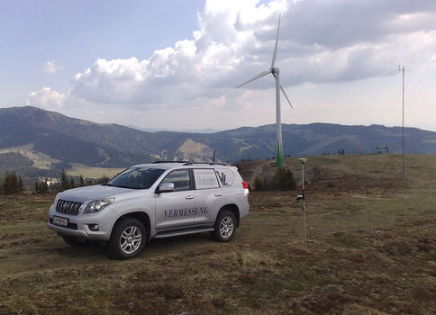 Weißes Geländefahrzeug mit der Aufschrift Vermessung steht auf einer offenen Wiese; daneben ein Vermessungsgerät. Im Hintergrund ragen Berge auf, und eine Windkraftanlage dreht sich unter einem wolkigen Himmel.