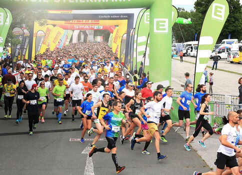 Eine große Gruppe Läufer startet einen Straßenlauf; grüne Startbögen mit Logos rahmen die Szene, während vorne einige in bunten Shirts sprinten und dahinter weitere Teilnehmer sowie Zuschauer folgen.