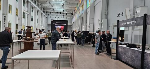 Große Messehalle mit Stehtischen und zahlreichen Besuchern. Links ein Hotel-Schild, rechts ein Verpflegungsstand 'Bon Appétit'. Im Hintergrund ein großer Bildschirm und weitere Ausstellerstände; helle Industrie-Beleuchtung und hohe Decken.