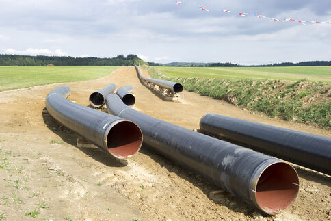 Mehrere große schwarze Metallrohre liegen in einem ausgegrabenen Graben auf offener Feldfläche – Teil einer Pipeline-Verlegung. Ringsum Felder, Absperrbänder und Baugerät im Hintergrund.