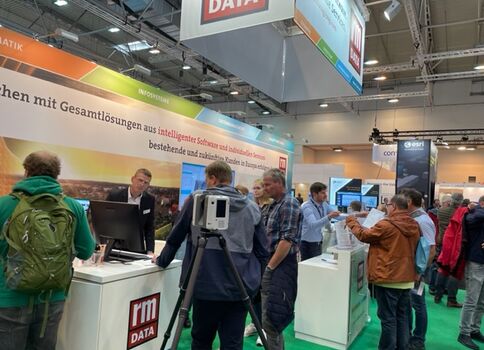 Mehrere Messebesucher stehen an einem Messestand der Firma rm DATA auf einer Technikmesse; weiße Theken mit Monitoren, Mitarbeiter führen Gespräche, im Hintergrund ein großes Banner mit DATA-Logo und weitere Messestände.