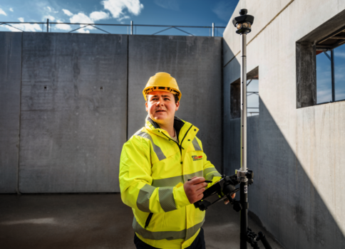 Ein Bauarbeiter in gelber Sicherheitsjacke und Helm bedient ein Vermessungsinstrument auf einer Baustelle. Er steht neben einer Betonwand, im Hintergrund der blaue Himmel; das Gerät ist auf einem Stativ montiert.