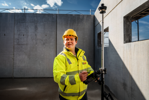 Ein Bauarbeiter in gelber Sicherheitsjacke und Helm bedient ein Vermessungsinstrument auf einer Baustelle. Er steht neben einer Betonwand, im Hintergrund der blaue Himmel; das Gerät ist auf einem Stativ montiert.