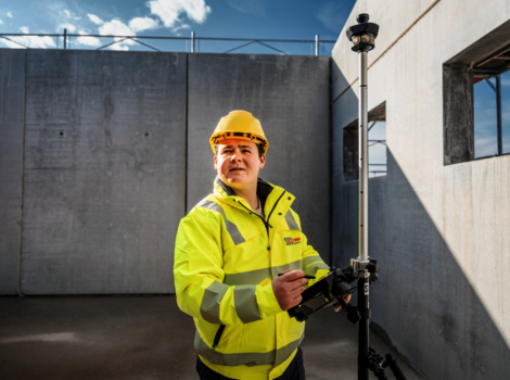 Ein Bauarbeiter in gelber Sicherheitsjacke und Helm bedient ein Vermessungsinstrument auf einer Baustelle. Er steht neben einer Betonwand, im Hintergrund der blaue Himmel; das Gerät ist auf einem Stativ montiert.
