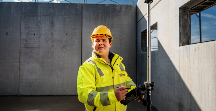 Ein Bauarbeiter in gelber Sicherheitsjacke und Helm bedient ein Vermessungsinstrument auf einer Baustelle. Er steht neben einer Betonwand, im Hintergrund der blaue Himmel; das Gerät ist auf einem Stativ montiert.