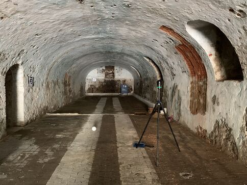 Alter Stein-Tunnel mit Gewölbedecke. Im Vordergrund steht ein Dreibein-Stativ mit Messgerät; der Tunnel führt in die Dunkelheit, rechts und links erkennbare Nischen.