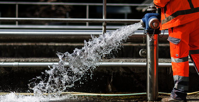 A worker wearing bright orange safety gear operates a high-pressure water jet from a nozzle on a metal pipe, spraying water across a wet ground.