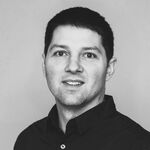 Porträt eines jungen Mannes mit kurzen dunklen Haaren, trägt schwarzes Hemd, schaut in die Kamera und lächelt leicht; grauer Hintergrund.