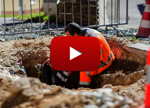 Ein Bauarbeiter in orange Warnkleidung kniet in einem offenen Graben und arbeitet an einer schwarzen Rohrleitung. Um ihn herum Baustellensicherung, Zäune und orange Verkehrskegel; im Hintergrund sind weitere Bauarbeiten sichtbar.