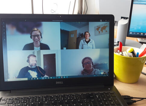 Auf einem Schreibtisch steht ein Dell-Laptop, dessen Bildschirm eine Videokonferenz mit vier Teilnehmern in separaten Fenstern zeigt.