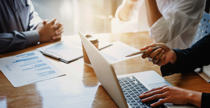 Mehrere Geschäftsleute sitzen um einen Konferenztisch. Ein Laptop ist geöffnet, daneben liegen Berichte mit Diagrammen. Eine Person hält einen Stift, die anderen diskutieren und schauen auf das Display.