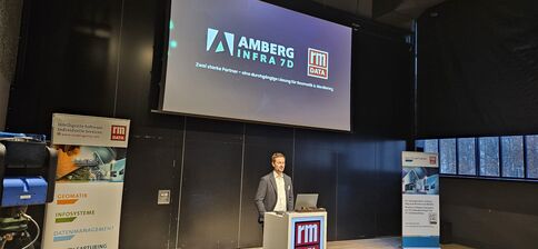 Ein Mann in Anzug steht hinter einem Rednerpult auf einer Bühne und hält eine Präsentation. Im Hintergrund zeigt eine Beamerprojektion Logos wie AMBERG INFRA 7D und rm DATA; Bühnenbanner an beiden Seiten der Bühne.