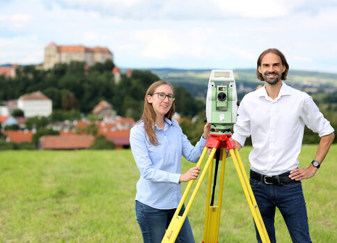 Zwei Vermessungstechniker stehen in einem Feld neben einem Vermessungsgerät auf einem Stativ; im Hintergrund eine ländliche Siedlung mit einer Burg auf dem Hügel.