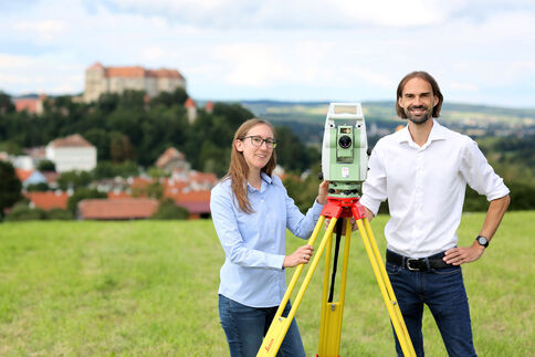 Zwei Vermessungstechniker stehen in einem Feld neben einem Vermessungsgerät auf einem Stativ; im Hintergrund eine ländliche Siedlung mit einer Burg auf dem Hügel.