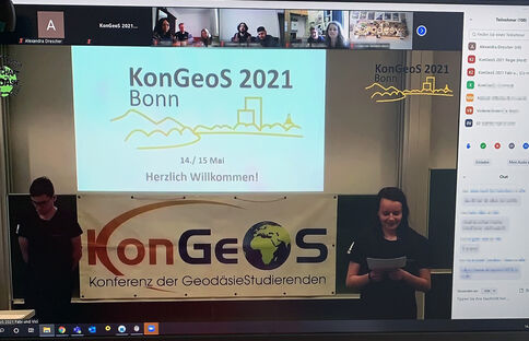 Zweiköpfiges Präsentationsteam steht auf der Bühne, während eine Projektion im Hintergrund „KonGeOS 2021 Bonn“ zeigt, mit „Herzlich Willkommen!“ und Datum 14./15 Mai. Vor der Bühne hängt ein Banner „KonGeOS – Konferenz der Geodäsie-Studierenden“.