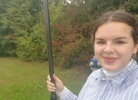 Eine junge Frau steht draußen im Park, hält einen langen schwarzen Stab mit einem orangefarbenen, runden Messgerät an der Spitze; sie lächelt in die Kamera.