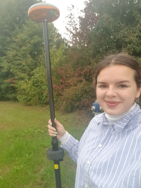 Eine junge Frau steht draußen im Park, hält einen langen schwarzen Stab mit einem orangefarbenen, runden Messgerät an der Spitze; sie lächelt in die Kamera.