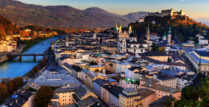 Luftaufnahme einer historischen Stadt am Fluss mit Brücken, Dom- und Kirchtürmen sowie einer Burg auf einem Hügel; dahinter erstrecken sich Berge.