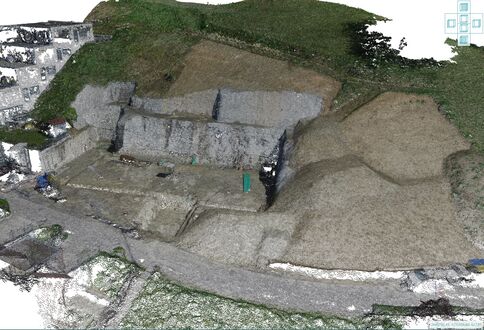 Terrasses en pierre taillées dans une colline, avec des murs et niveaux visibles; zone centrale dégagée; tas de gravier à droite et chemin pavé à gauche, entouré de verdure.