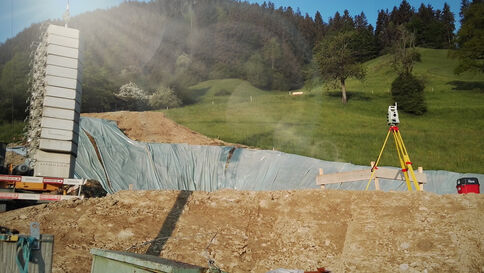 Eine Baustelle mit einer großen Erdmulde, teilweise von blauer Plane bedeckt. Links steht ein vertikales Bauwerk, rechts ein Vermessungsgerät auf einem Dreibein. Im Hintergrund grüne Hügel und Wald, sonniges Wetter.