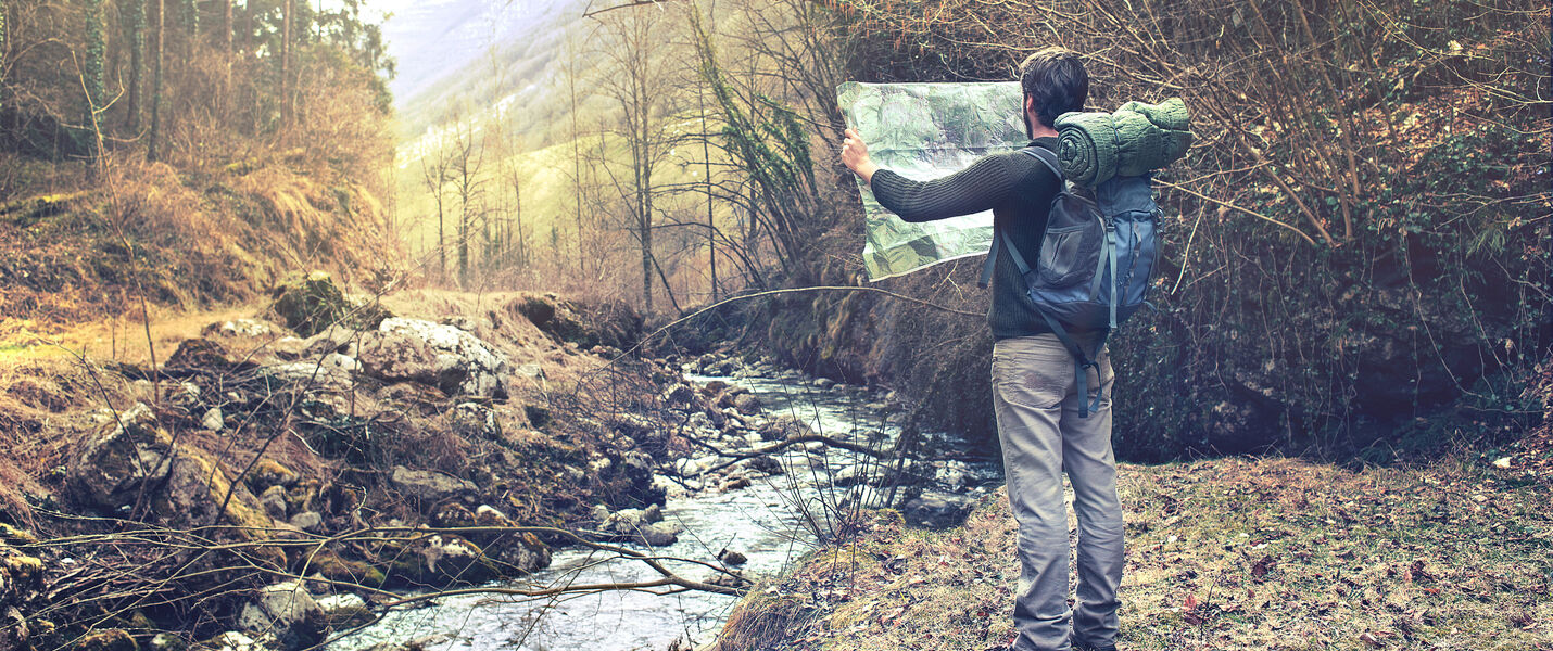 Ein Mann mit Rucksack steht am Ufer eines kleinen Flusses im Wald und hält eine Karte ausgestreckt, als Orientierungshilfe. Hinter ihm sehen wir Bäume, den Flusslauf und entfernte Berge in einer herbstlichen Landschaft.