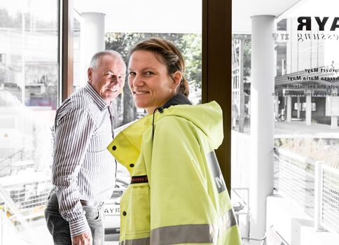 Zwei Personen stehen im hellen Eingangsbereich eines Gebäudes. Im Vordergrund lächelt eine Frau in einer neon-gelben Sicherheitsjacke mit reflektierenden Streifen in die Kamera; im Hintergrund steht ein Mann in gestreiftem Hemd. Große Glasfronten und Türen zeigen die Außenwelt.