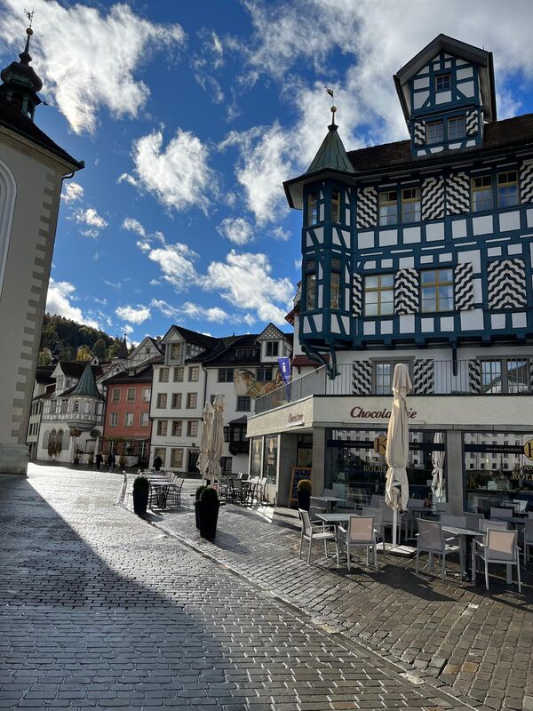 Malerischer europäischer Stadtplatz mit gepflasterter Oberfläche, draußen Café-Tische unter Schirmen. Im Hintergrund ein blau-weißes Fachwerkhaus und weitere bunte Gebäude; der klare Himmel mit Wolken und einige Passanten, die durch den Platz schlendern.