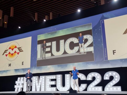 Eine Konferenzbühne mit drei großen Bildschirmen; in der Mitte stehen zwei Redner vor einer EUC22-Anzeige. Vorne leuchtet ein großer Hashtag "#IMEUC22".