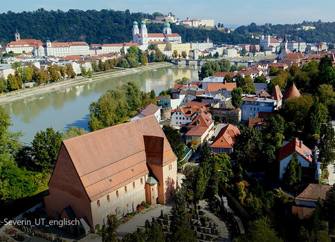 Vue aérienne d'une ville près d'une rivière: au premier plan se dresse une grande bâtisse en pierre au toit rouge entourée de maisons et d'arbres; plus loin, la rivière serpente et des collines couvertes de bâtiments et de verdure dominent l'horizon.