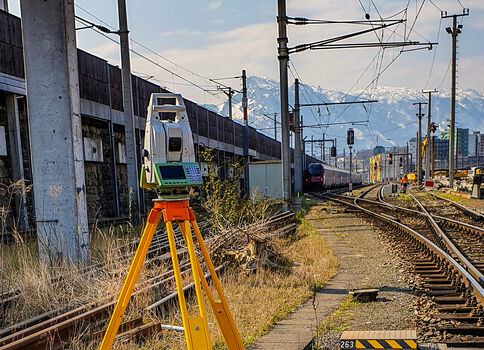 Vermessungsgerät auf einem gelben Dreibeinstativ steht neben einer Eisenbahnschiene; dahinter fahren Züge, Oberleitungsmasten ragen in den Himmel. Die Szene zeigt eine Bahnstrecke mit Weichen, Gebäuden und einer Bergkette im Hintergrund.