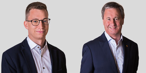 Zwei Männer in Anzügen posieren vor einem hellgrauen Hintergrund; links mit Brille, rechts mit grauen Haaren. Beide schauen freundlich in die Kamera.