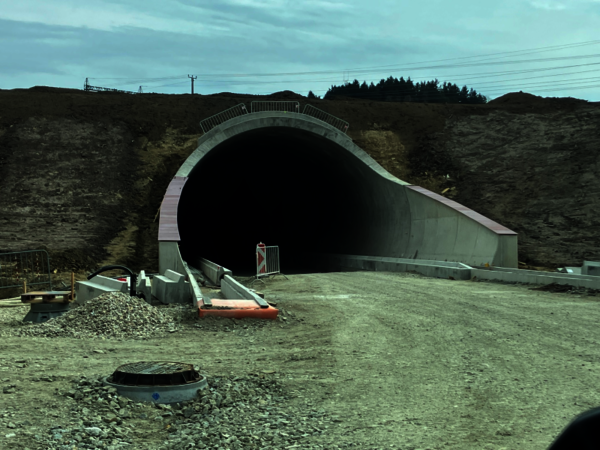 Große Tunnelbaustelle: Ein dunkler, weit ins Bergprofil gehender Tunneldurchgang mit Betonbogen; Umgeben von Bauzäunen, Barrieren und Schutt. Vor dem Eingang liegt ein runder Deckel. Der Weg ist unbefestigt; wolkiger Himmel über einer hügeligen Landschaft.