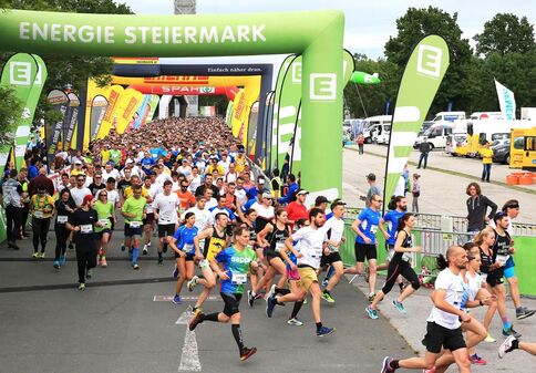 Viele Läuferinnen und Läufer starten ein Straßenrennen unter einem großen grünen Startbogen mit der Aufschrift ENERGIE STEIERMARK. Im Hintergrund befinden sich weitere Teilnehmer, Sponsorfahnen, Werbebanner sowie Zelte und Veranstaltungsfahrzeuge.