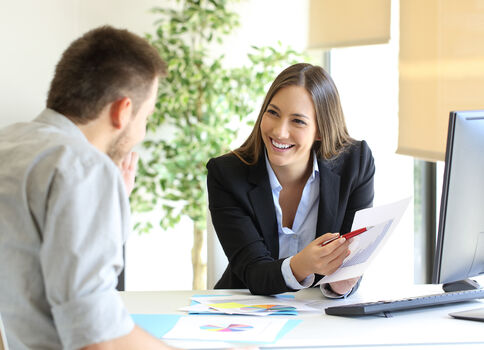 Eine berufliche Besprechung: Eine lächelnde Frau in Blazer reicht einem Mann Unterlagen über den Schreibtisch; Computer und eine Pflanze im Büro.