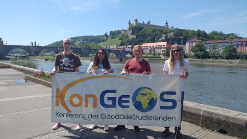Vier Personen stehen am Uferweg eines Flusses, halten ein Banner der Konferenz der Geodäsie-Studierenden mit Globus-Logo. Im Hintergrund sind eine Brücke, eine Stadt mit roten Gebäuden und eine Burg auf dem Hügel zu sehen.