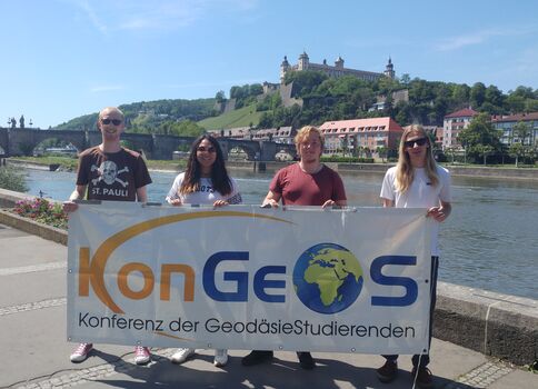Vier Personen stehen am Uferweg eines Flusses, halten ein Banner der Konferenz der Geodäsie-Studierenden mit Globus-Logo. Im Hintergrund sind eine Brücke, eine Stadt mit roten Gebäuden und eine Burg auf dem Hügel zu sehen.