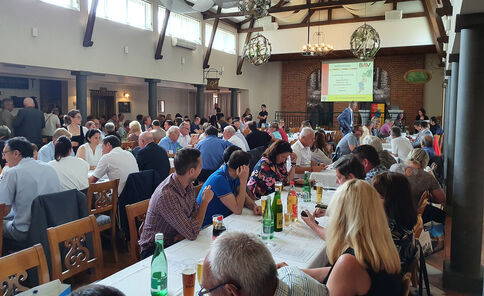 Großer Veranstaltungsraum mit vielen Gästen, die an langen Tischen speisen und trinken. Im Hintergrund eine Bühne mit Vortrag und projizierter Präsentation; eine Person steht am Rednerpult. Die Atmosphäre wirkt wie ein gemeinsames Essen oder Fest.
