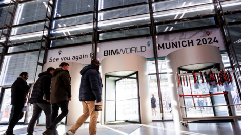 Innenaufnahme eines modernen Messegebäudes mit Glasfassade. Mehrere Personen gehen durch einen gläsernen Eingang, vor dem Banner 'BIM WORLD MUNICH 2024' hängt; rechts ein Stand mit Lanyards.